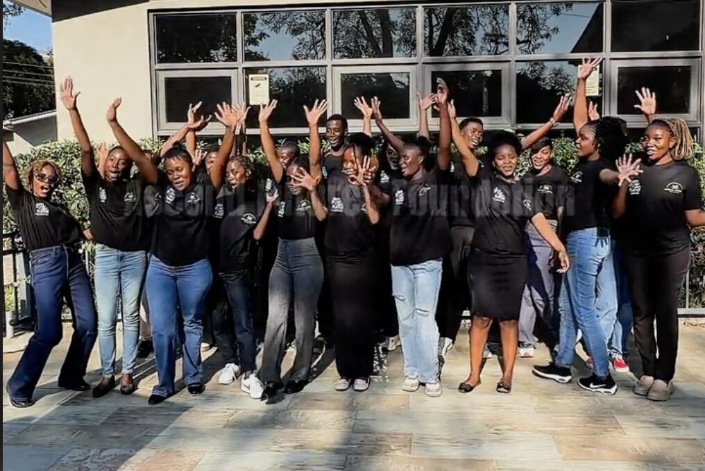 A large group of young women in matching black T-shirts cheering with their arms raised outside a building