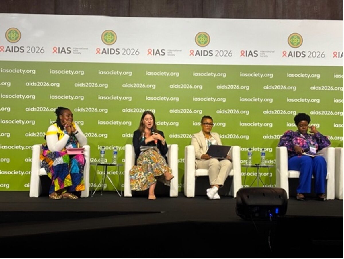 Kate Segal speaking into a microphone on a panel of four women seated on stage at AIDS 2026